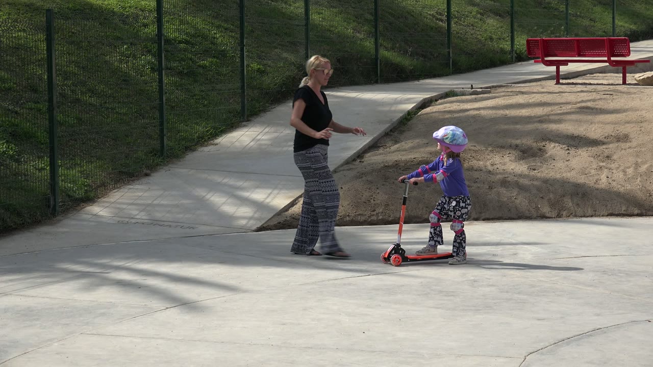 In Los Cabos, Mexico, a woman watches a small helmeted child standing on a red-and-black scooter on a sunny concrete path beside sand and a red bench.
