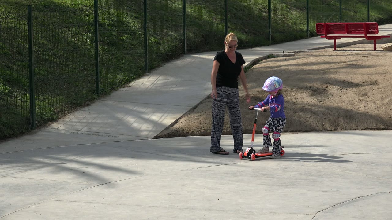 The woman faces the child as the helmeted child holds the scooter handlebar on the concrete, with a sand patch, red bench, fence, and grass behind them in Los Cabos, Mexico.