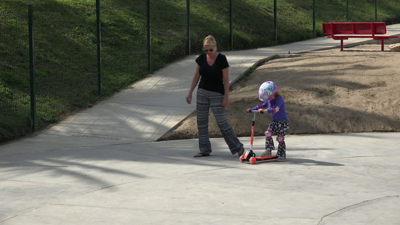 Child rides a scooter while a woman walks close by on a paved path.