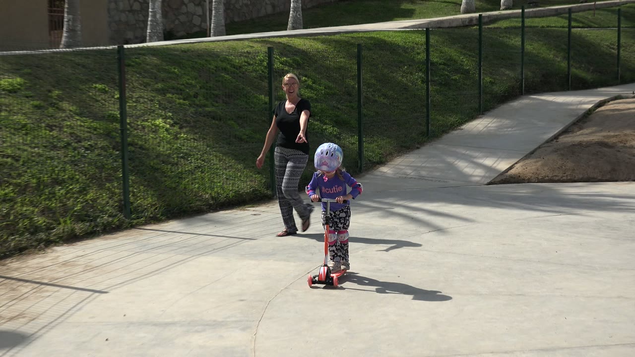 Young girl riding a red scooter with an adult woman walking behind her on a park path.