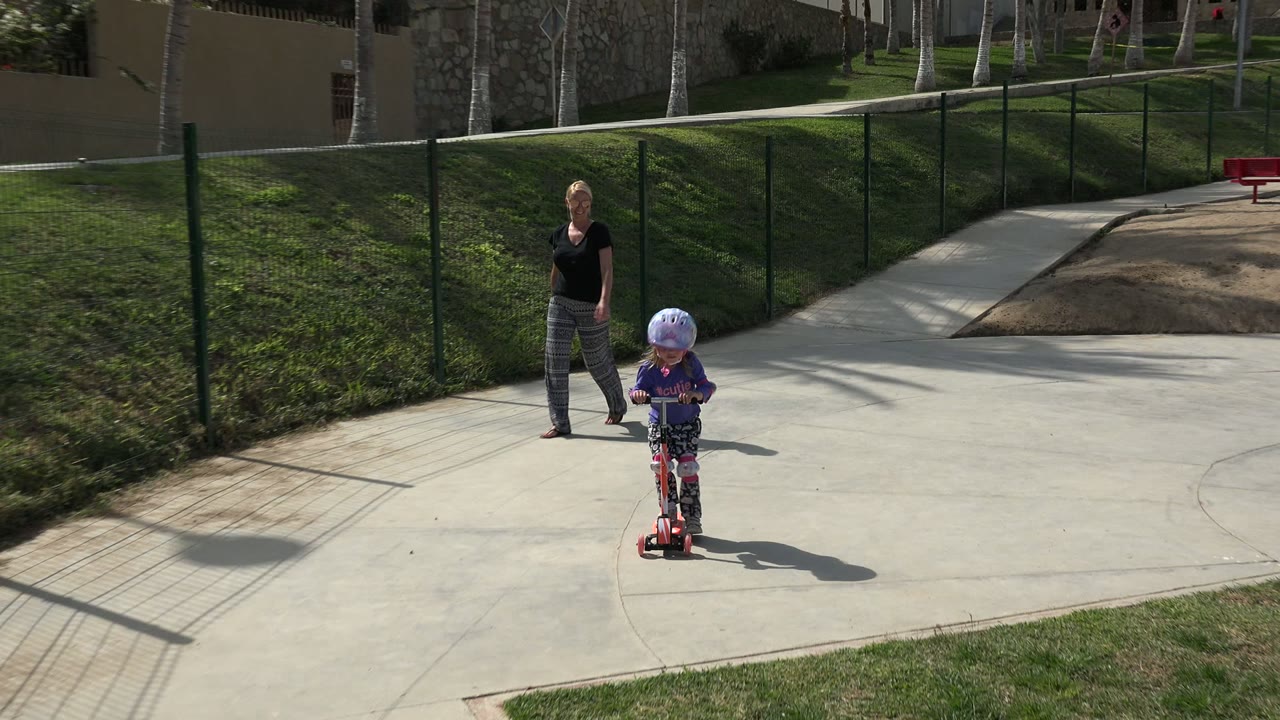 The child on the red scooter moves along the curved paved path in Los Cabos, Mexico, while the adult woman stays several steps behind near the fenced grass bank.