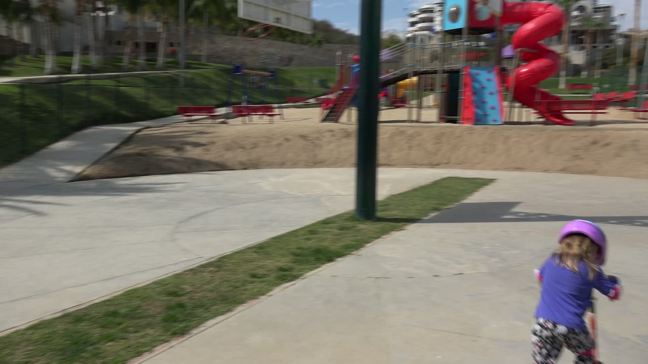 Child in a purple helmet riding a red scooter toward a playground.