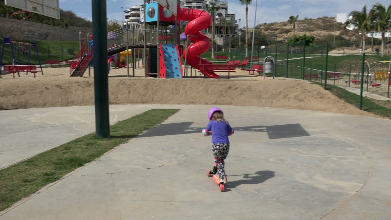 The child rides farther away on the red scooter across pale concrete in Los Cabos, with a foreground pole, playground fencing, palms, and dry hills behind.