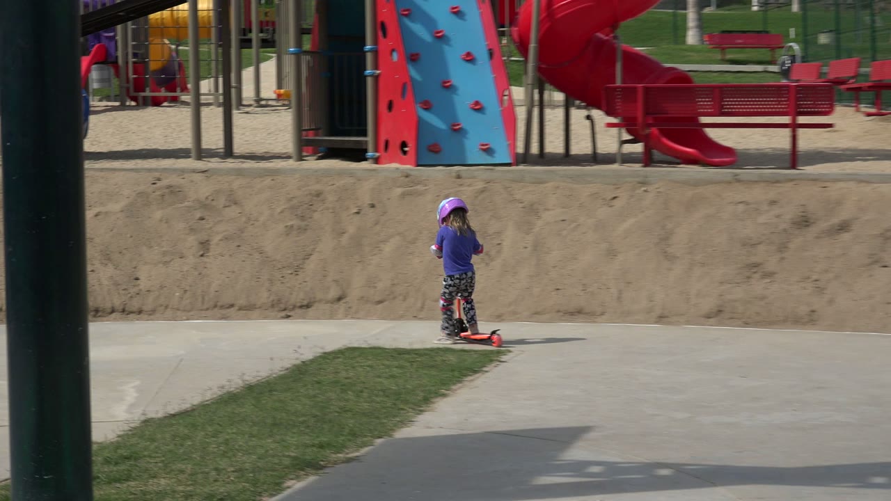 The child continues across the concrete in Los Cabos, Mexico, passing between a grass strip in front and red benches and playground structures behind.