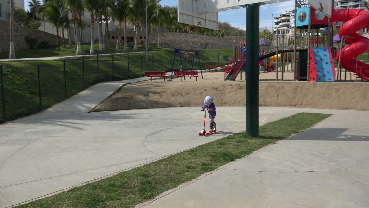 The child moves farther across the paved park path in Los Cabos, with a red bench, swings, chain-link fence, grass slope, and playground slide in the background.