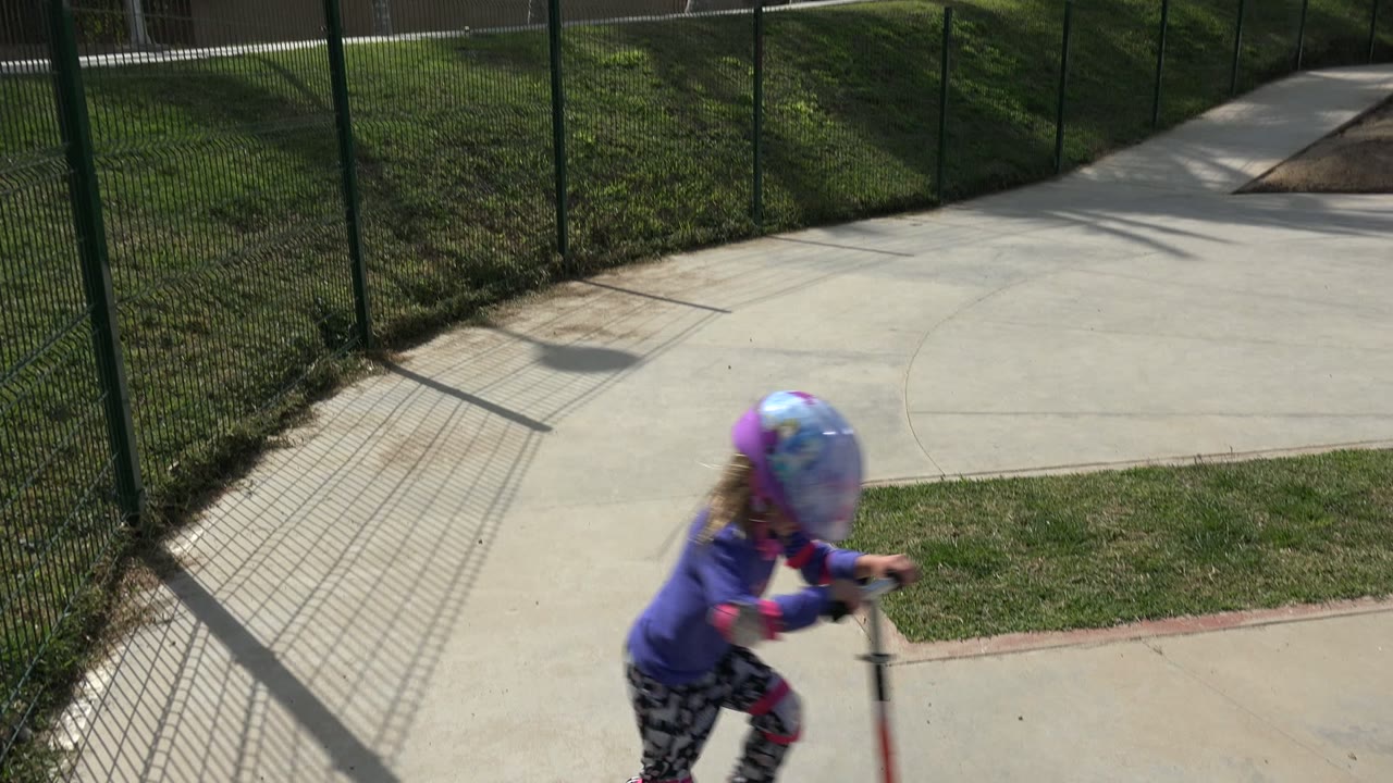 Young girl in a purple helmet riding a scooter beside a fence.