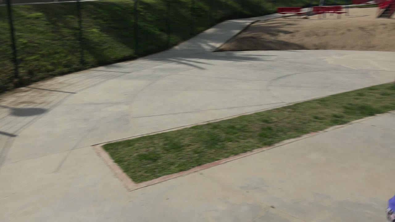 Concrete walkways curve through a Los Cabos park with grass strips, fenced edges, sand play areas, and the playground partially visible in the distance, with no visible people.