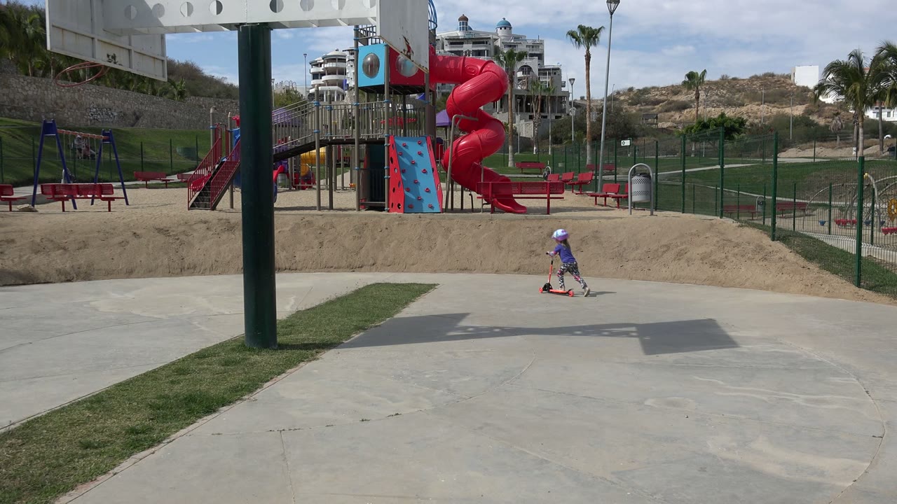 Helmeted child riding a scooter near a playground slide