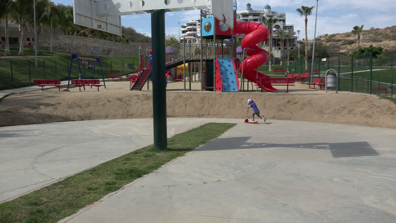 The child continues across the concrete path with the playground, fencing, palms, and hillside buildings behind them in Los Cabos, Mexico.