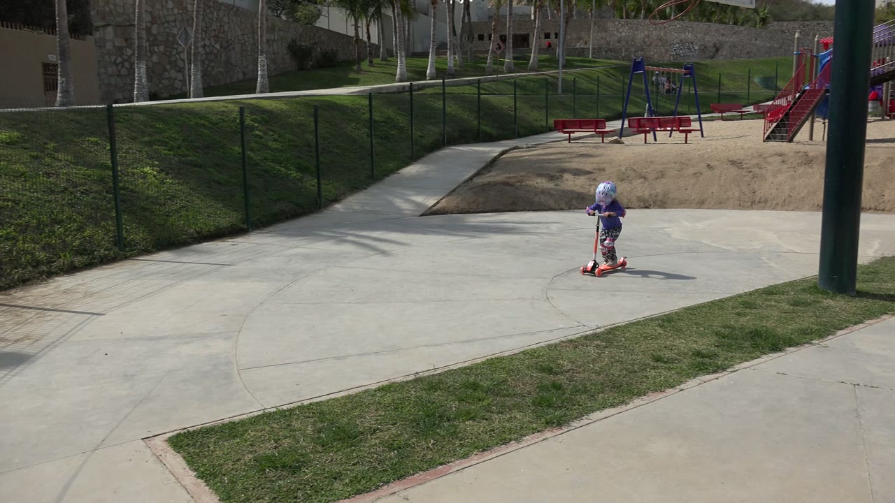 Child on an orange scooter rides along a concrete park path.