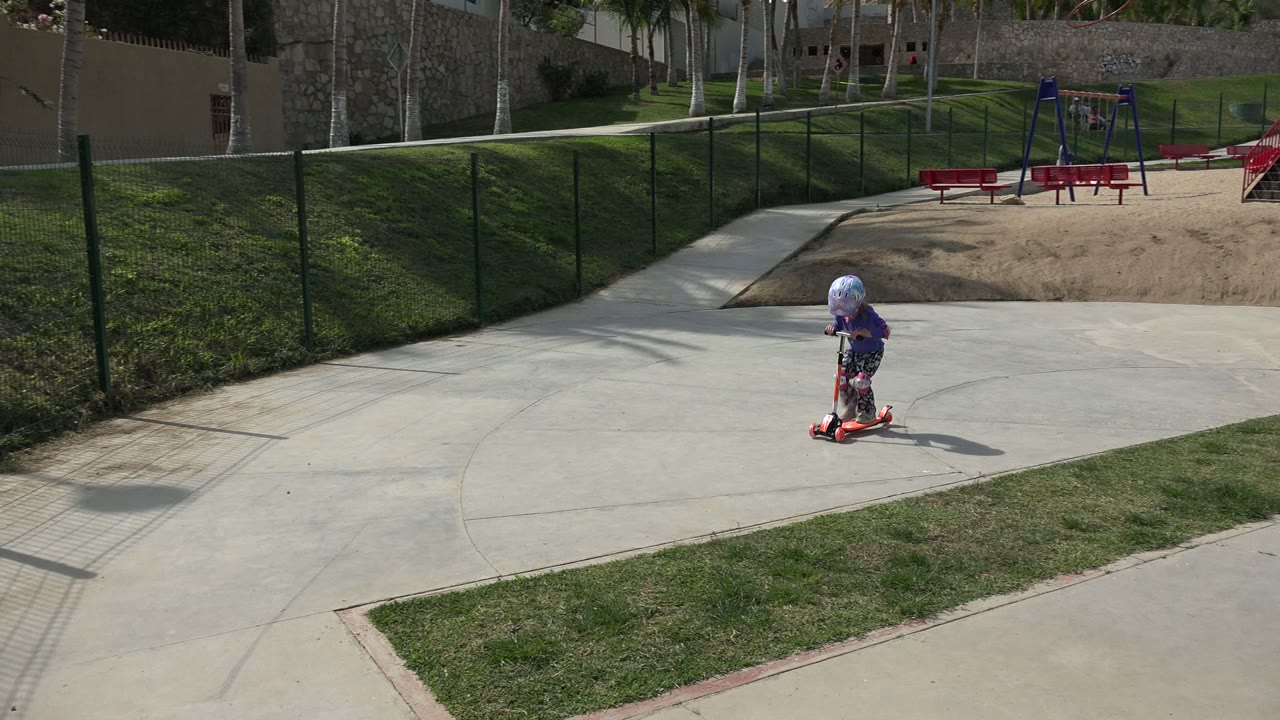 The child continues across the pale concrete walkway in Los Cabos, leaning over the scooter handlebars with a grassy slope and black fence in the background.