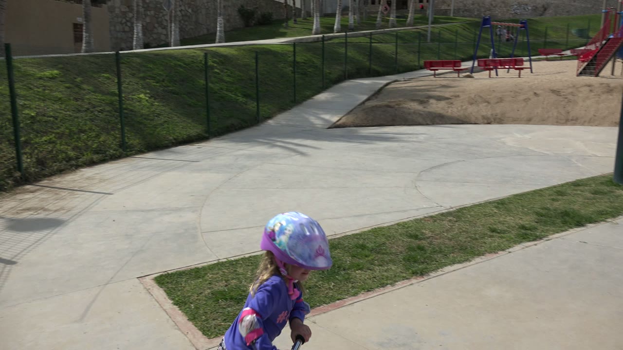 The young girl moves farther into frame with scooter handlebars visible on a wide concrete path in Los Cabos, Mexico, backed by grass, fencing, benches, and a small play structure.