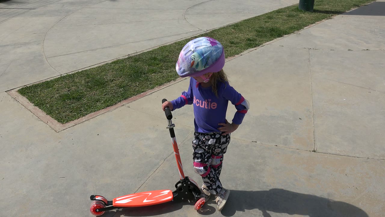 The child looks down near the scooter handlebar, wearing a colorful helmet and pads on a concrete playground area in Los Cabos, Mexico.