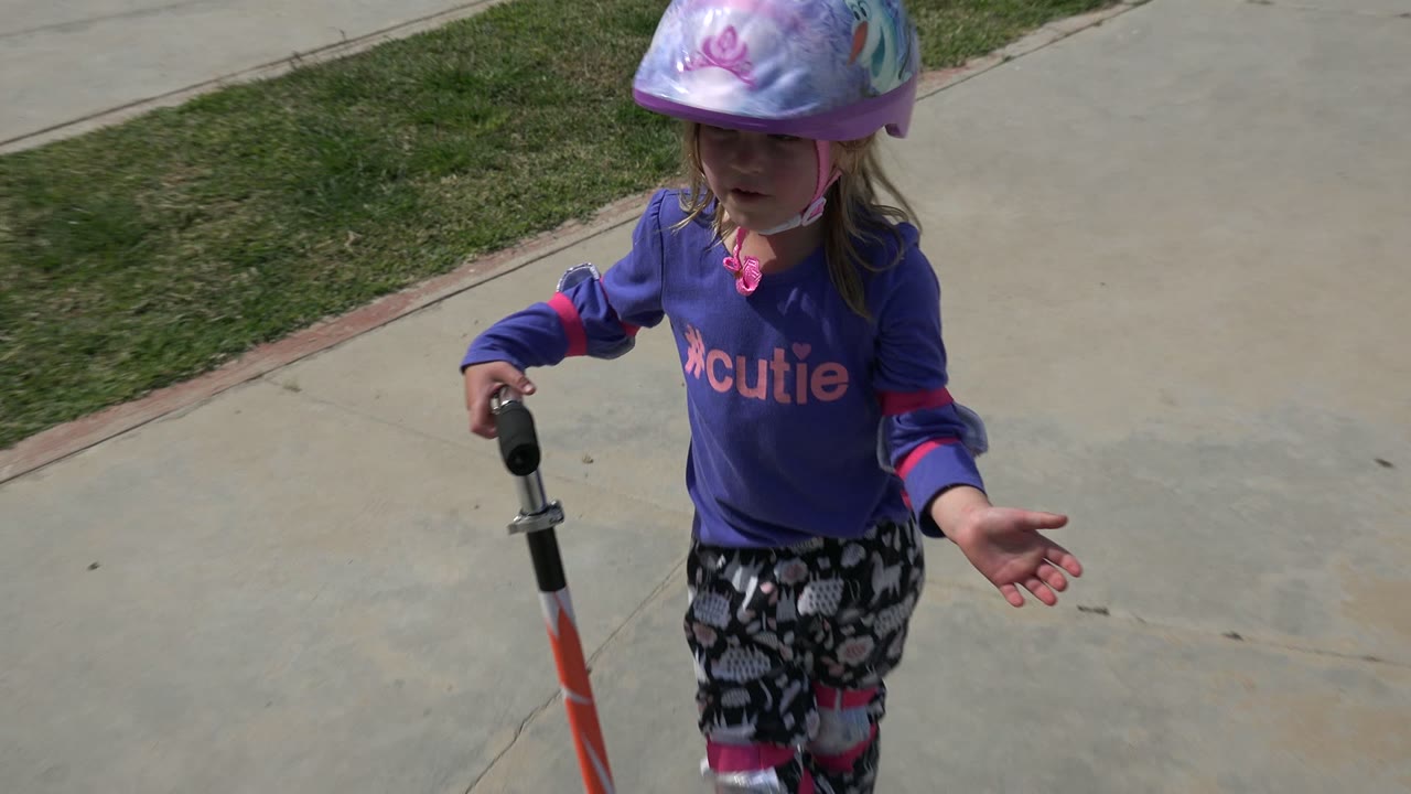 A young girl in Los Cabos, Mexico wears a purple character helmet and purple shirt while standing with a scooter on a concrete path beside grass.