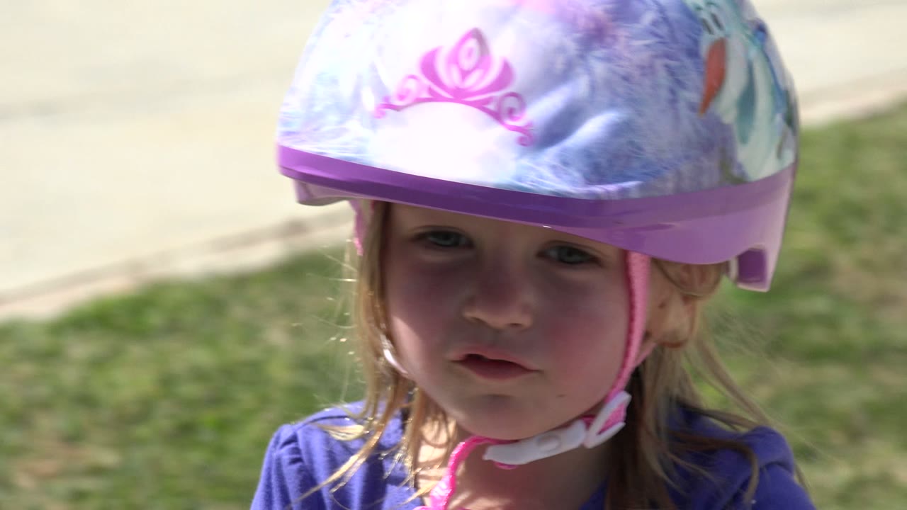 Close-up in Los Cabos, Mexico of a blonde child facing slightly downward in a purple illustrated safety helmet, with grass and sidewalk behind her.