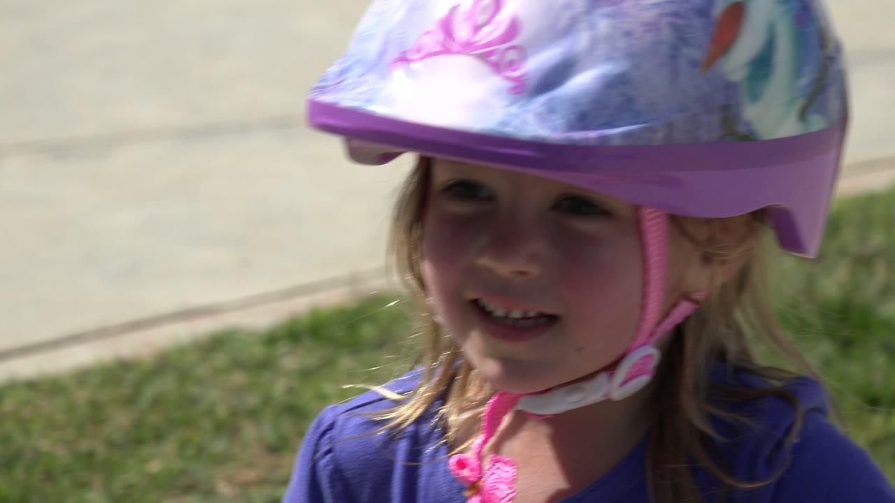 Close-up in Los Cabos, Mexico of the helmeted blonde child smiling toward the camera, with a sunlit sidewalk and lawn in the background.