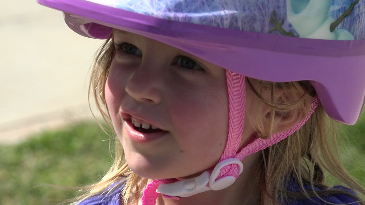 A young blonde girl in a purple helmet looks to the side while standing near a sidewalk and grass in Los Cabos, Mexico.