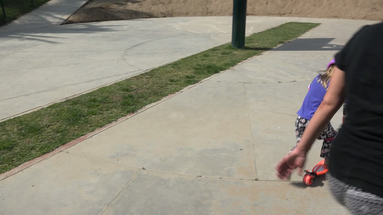 Adult walking beside a young girl on a scooter in a concrete play area.