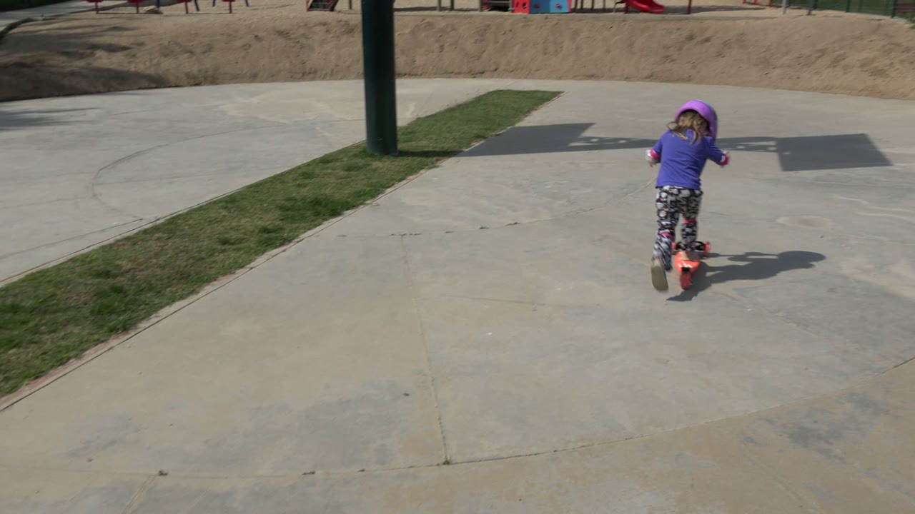 In Los Cabos, Mexico, the young girl rides the scooter away from camera across a wide concrete area, with grass strips, sandy ground, and playground equipment in the background.