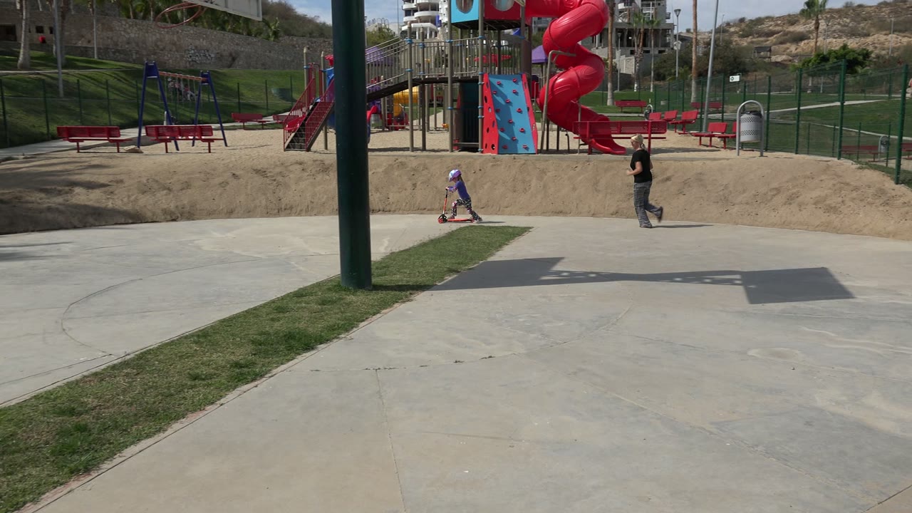 At 0:00 in Los Cabos, Mexico, a helmeted child rides a scooter on a concrete park path while an adult walks nearby beside a playground and sandy play area.
