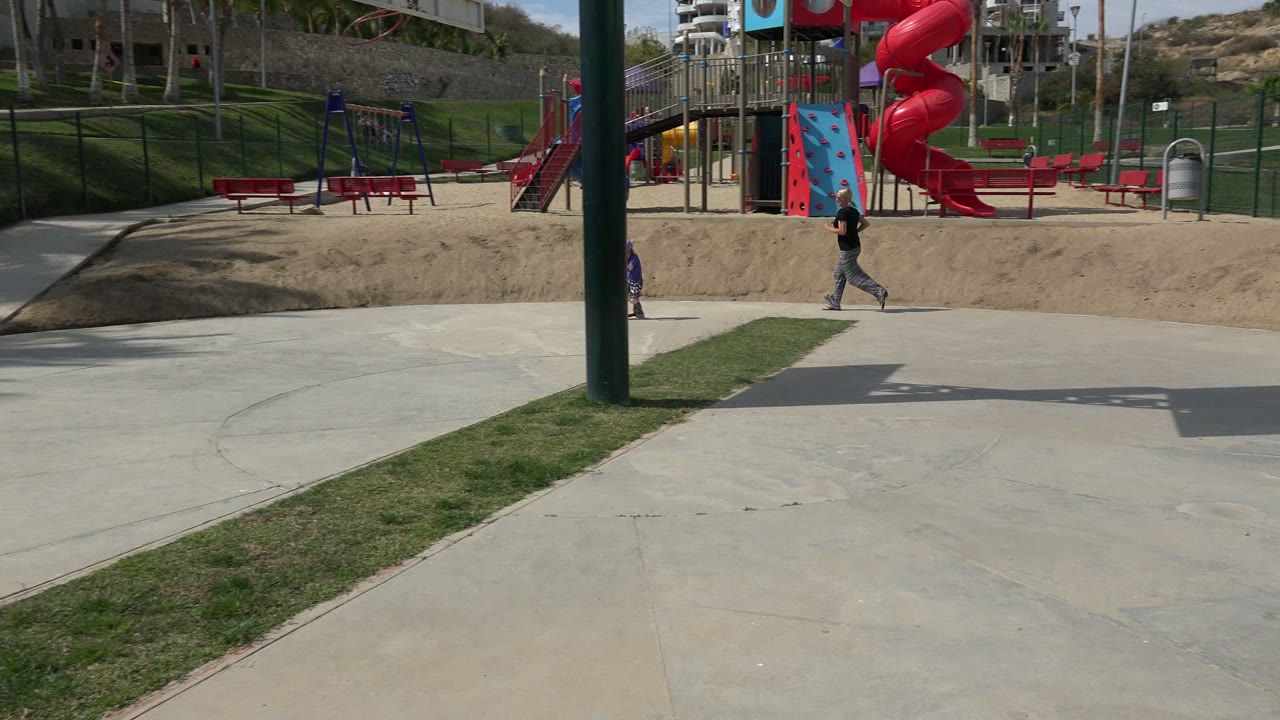 At 0:01 in Los Cabos, Mexico, the child on the scooter has moved left of frame while the adult follows across the path near red playground structures and benches.