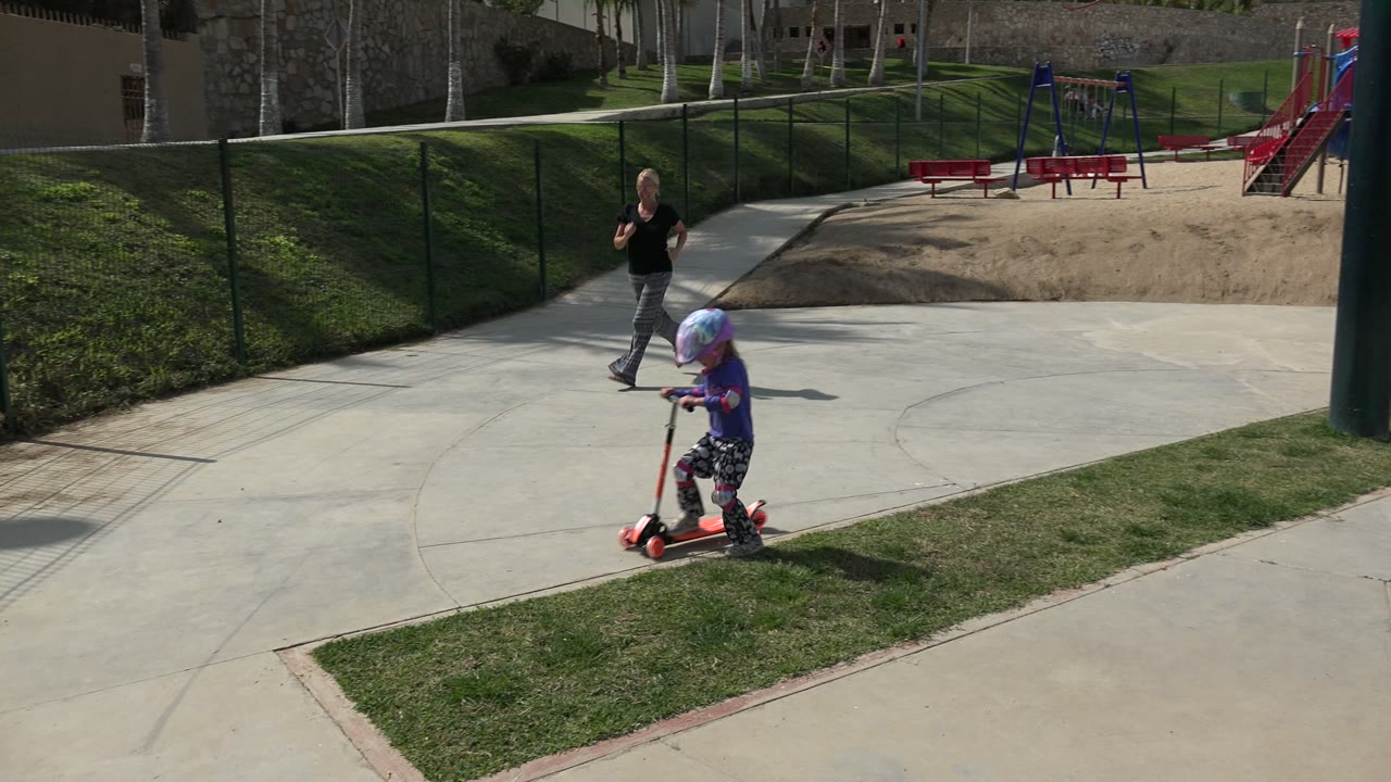 At the start of the Los Cabos clip, a helmeted child rides a red scooter across a sunlit paved area as an adult woman follows behind near fencing and playground equipment.