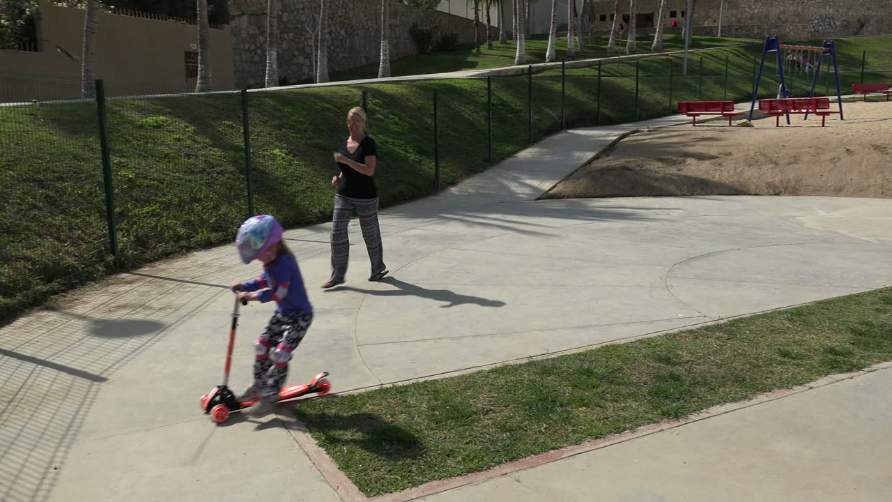 A helmeted child moves farther across the Los Cabos paved play area on a red scooter while an adult woman stands in the midground near the fence and curved sidewalk.