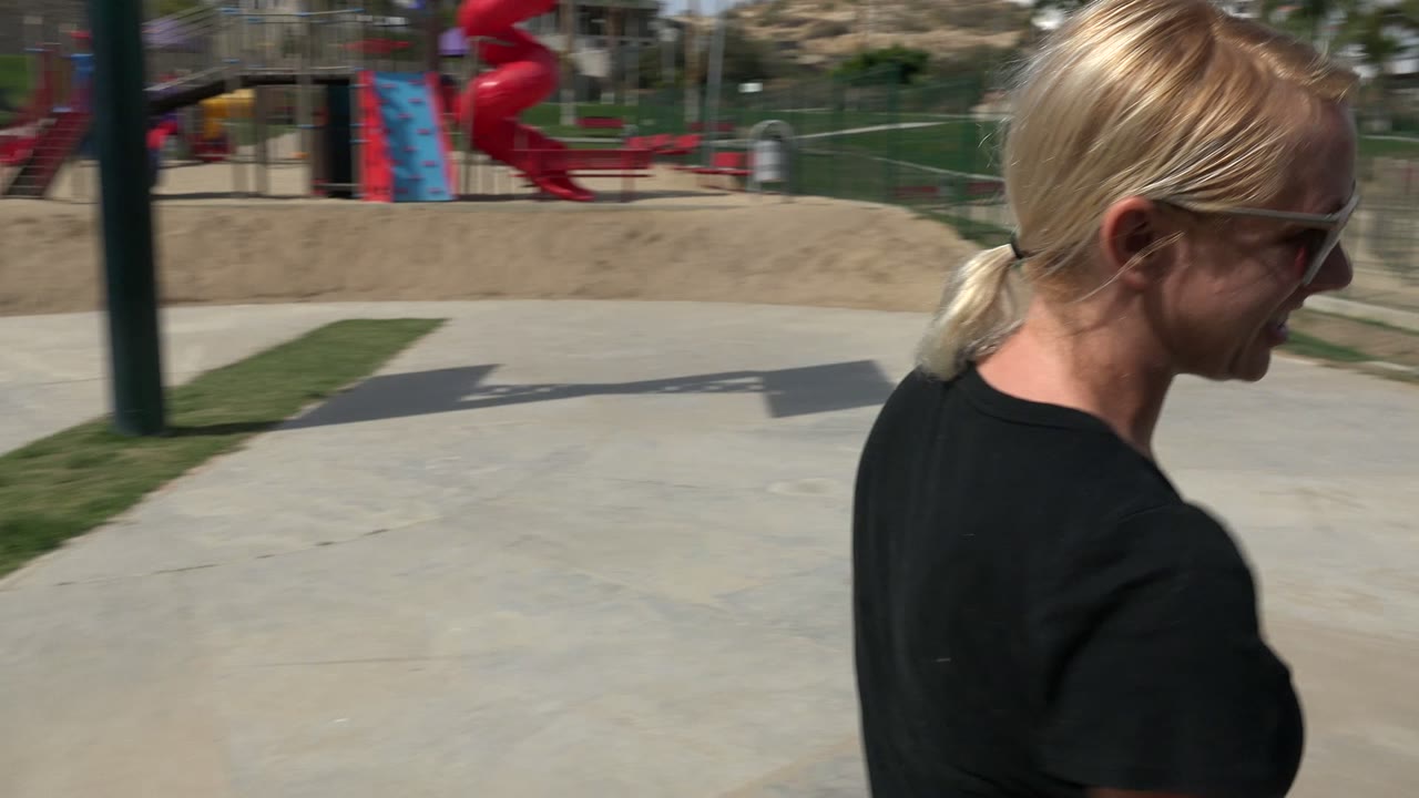 Blonde woman in sunglasses walks through a sunny fenced park area in Los Cabos, Mexico, with playground equipment and palms in the background.