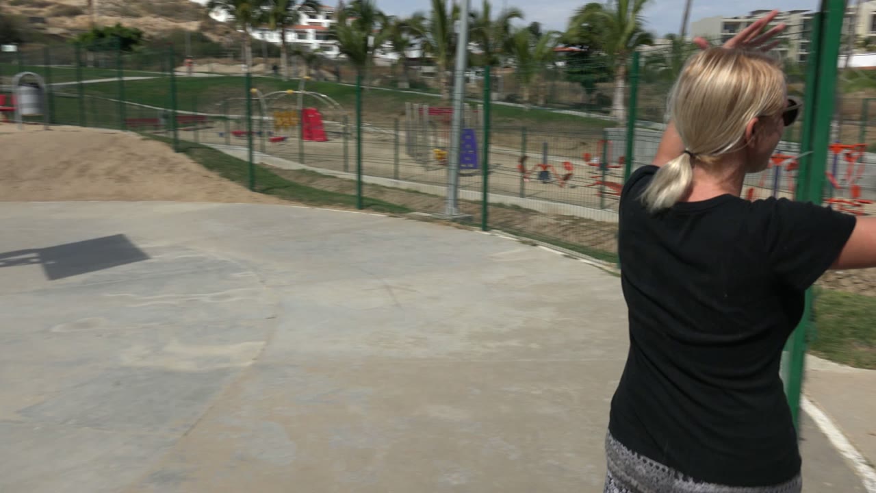 The woman is seen from behind near a green fence in a sunny Los Cabos park, with red and blue playground structures beyond the path.