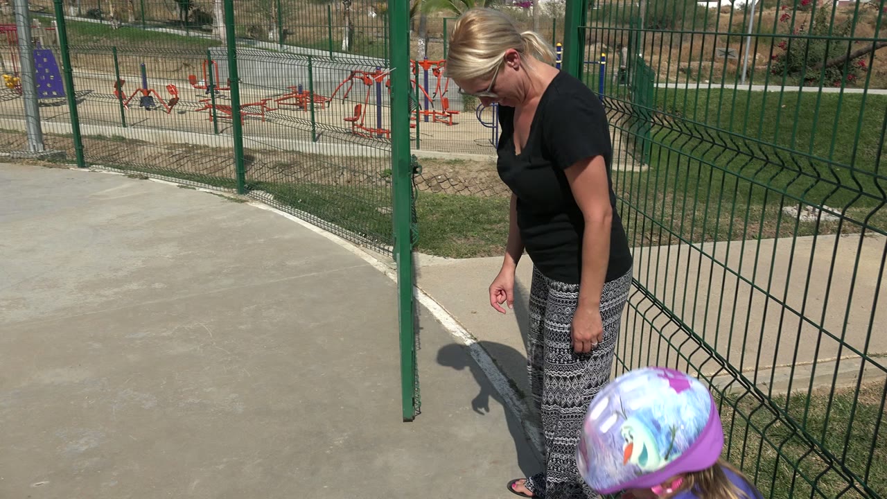 The camera view holds on the concrete walkway and green playground fence in Los Cabos, Mexico, with the woman mostly cropped at the right edge.