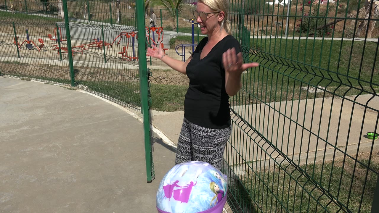 At a Los Cabos playground, a woman in sunglasses gestures with both hands while a helmeted young girl is partly visible at the bottom of the frame.