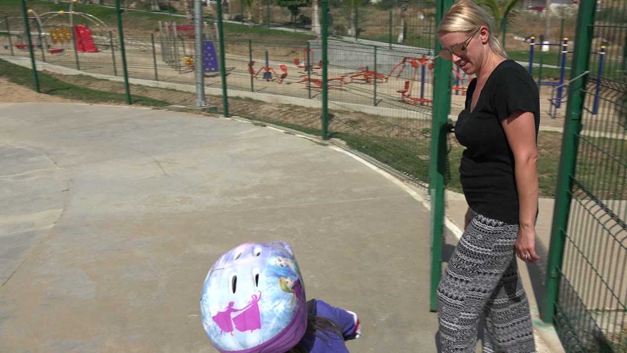 Woman standing beside a helmeted child at a concrete skate park.