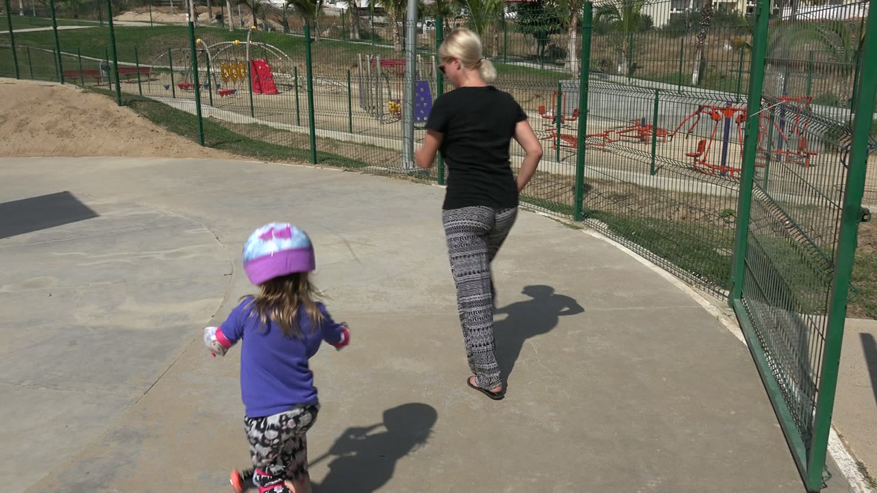Young girl on a scooter following an adult woman in a fenced park.