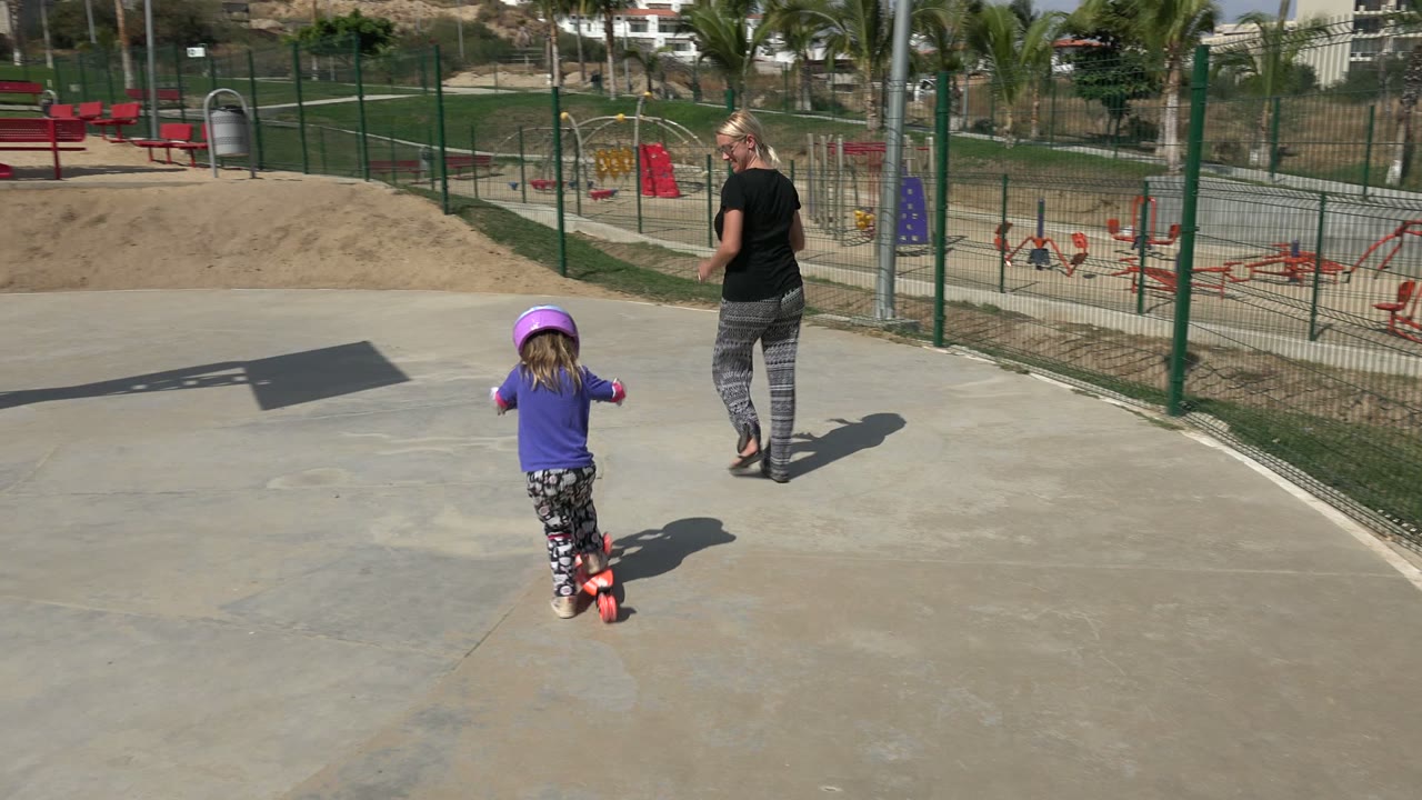 The child continues on the red scooter toward the adult woman in the Los Cabos park, with a fenced play area, red equipment, grass, and palm trees in the background.