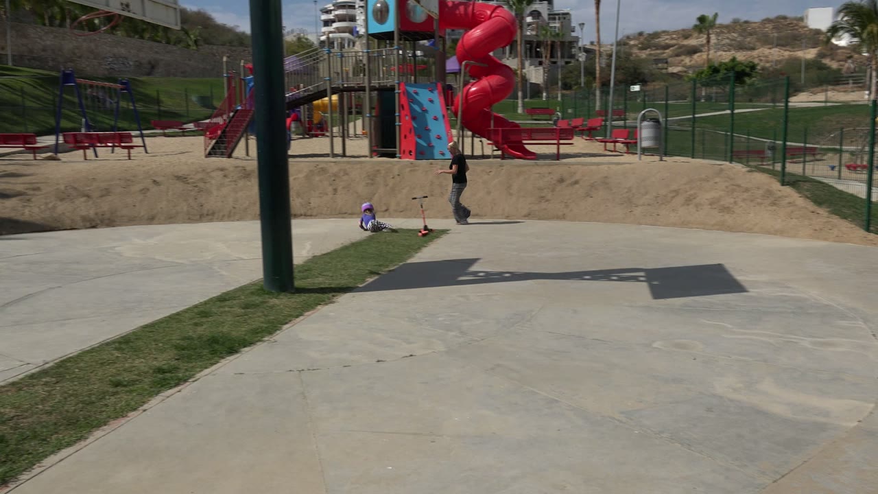 Wide view of a child near a scooter at a skate park with an adult nearby.