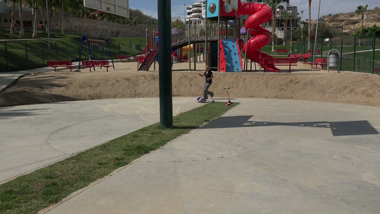 In Los Cabos, Mexico, the helmeted child is down near the scooter beside a narrow grass strip while the adult approaches from the right side of the concrete path.