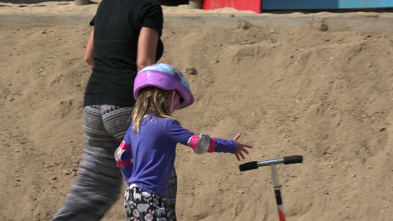 In Los Cabos, Mexico, a young girl in a purple helmet reaches toward an orange scooter while an adult woman stands partly cropped beside her near a sandy bank.