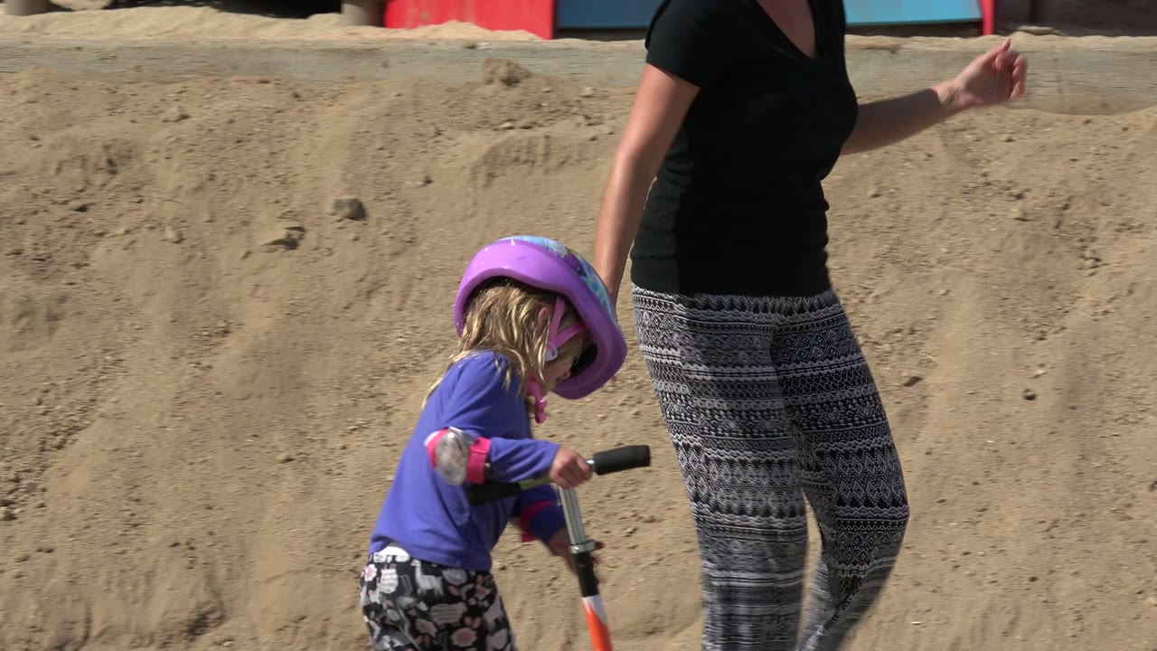 In Los Cabos, Mexico, the helmeted girl holds the orange scooter handlebar as an adult woman walks next to her against the sandy playground edge.