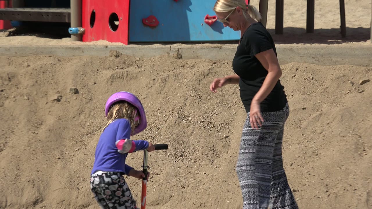 Girl with orange scooter near a sandy bank as an adult stands to the right.