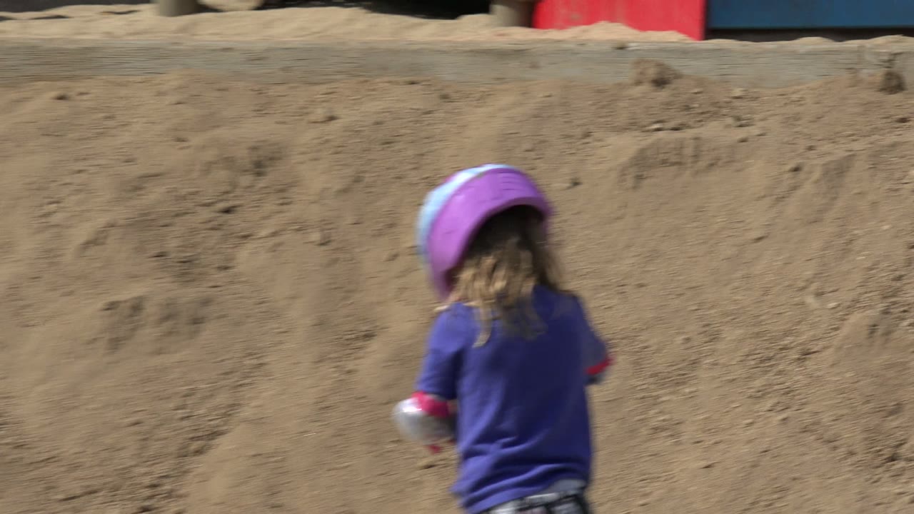 Child in purple helmet riding a scooter near a sandy hillside.