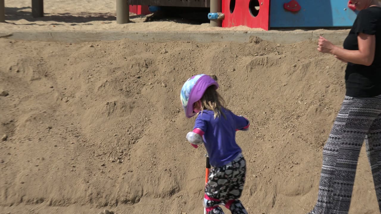 Child in a helmet rides a red scooter near an adult on a sunlit playground path.