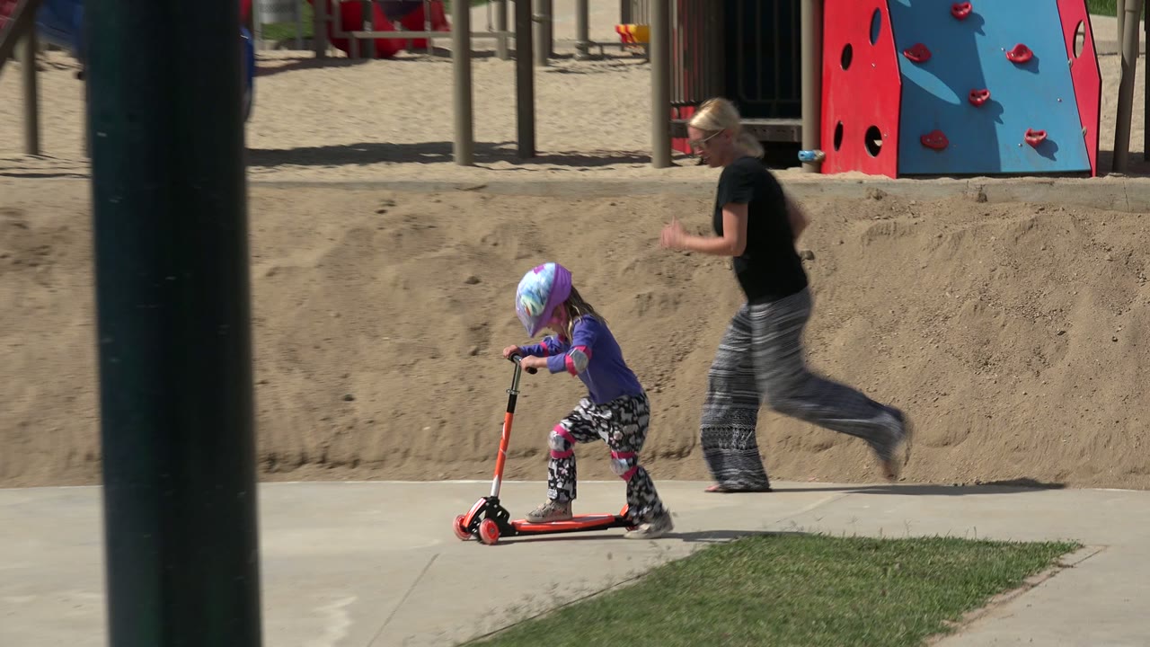 In Los Cabos, Mexico, the child rides the red scooter across the concrete path as an adult woman runs behind, with red benches and climbing play equipment in the background.