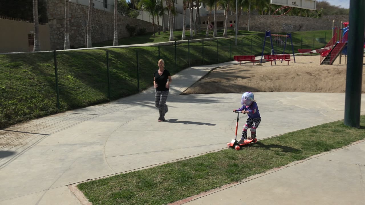 In Los Cabos, Mexico, a woman walks on a concrete path toward a helmeted little girl on a red scooter beside a grass strip and fenced playground.