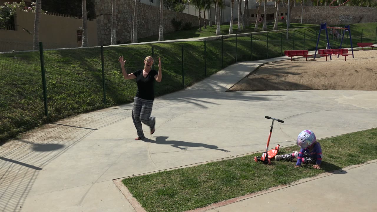 In Los Cabos, Mexico, the mother lifts her hands on the path as the little girl sits or falls on the grass next to the red scooter near the fenced playground.