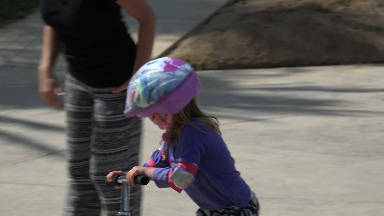 Child in helmet riding a scooter beside an adult on a paved path.