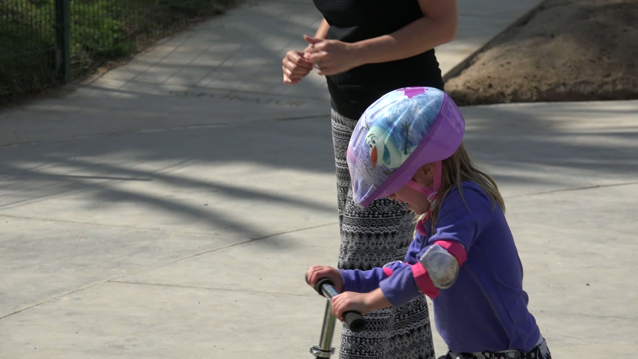 A young girl in a colorful helmet and purple top rides her scooter through the Los Cabos park path while the adult is partly visible beside her.