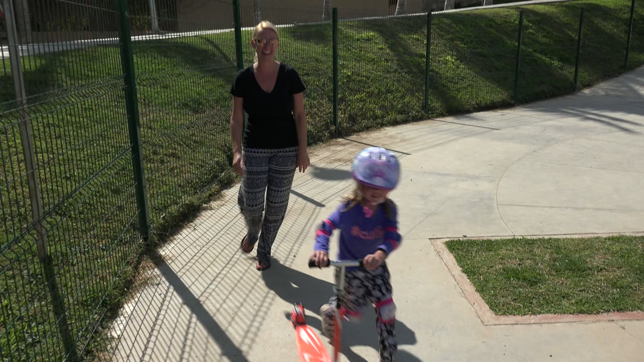 In Los Cabos, a blonde mother in sunglasses walks behind a helmeted child on a scooter beside a tall green fence and sunlit pavement.