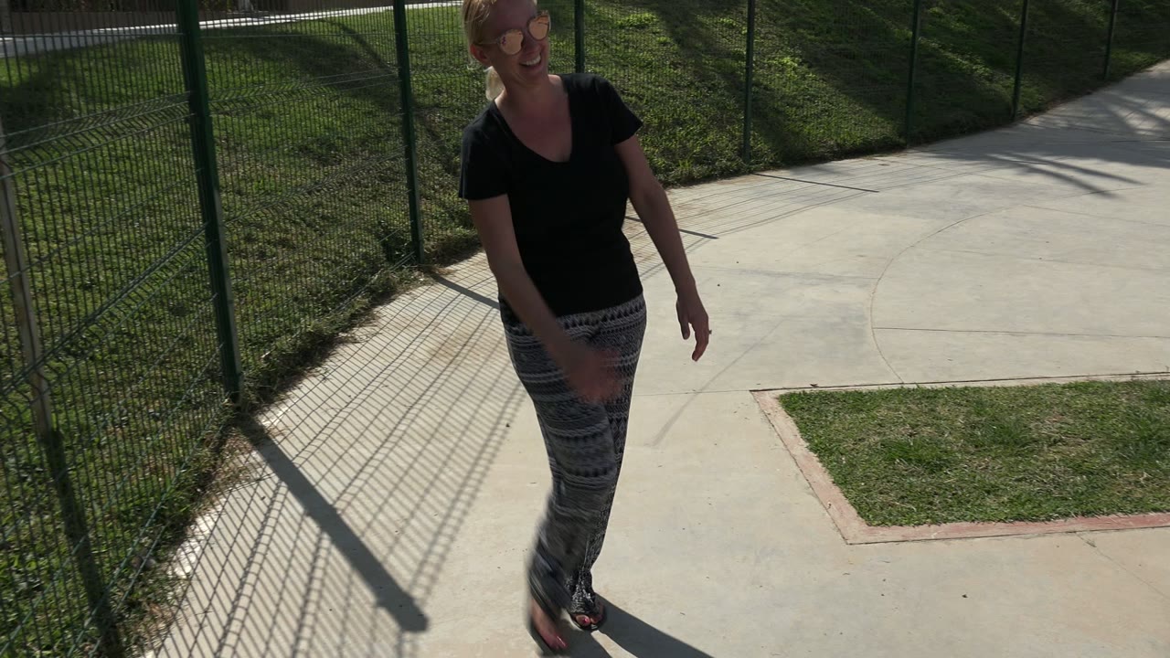 In Los Cabos, the mother smiles while walking on a concrete sidewalk next to a green fence, with grass and hard fence shadows across the path.