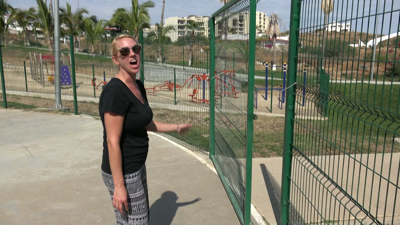 Woman in sunglasses stands beside a green metal fence at a Los Cabos park, with playground equipment and palm trees visible behind her.