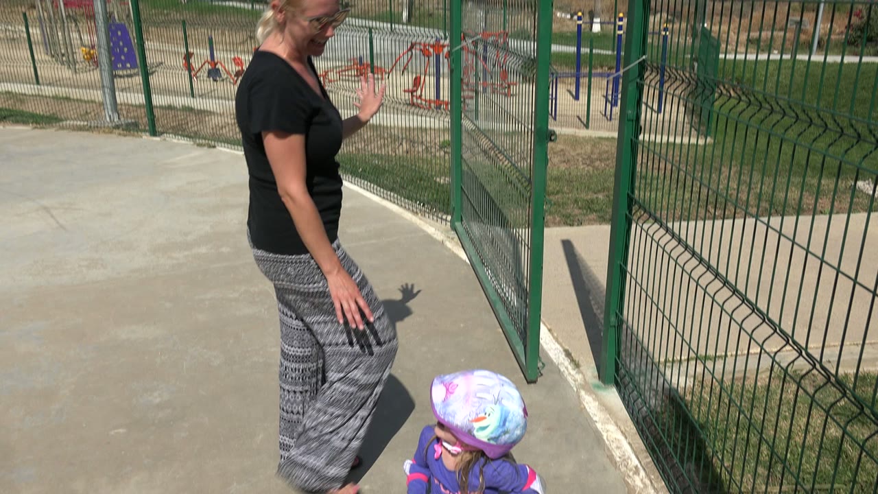 Woman moves along the concrete path beside a small child in a purple helmet near the green fence at the Los Cabos playground.